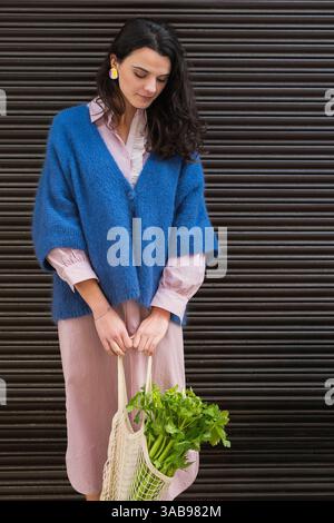 Une femme portant un cardigan bleu et une robe rayée tient un sac en maille rempli de verts frais. Ses boucles d'oreilles colorées ajoutent une touche vibrante, contrastant wi Banque D'Images