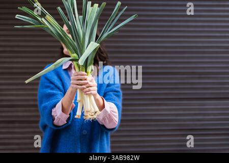 Une femme en pull bleu tient un paquet de légumes frais, cachant son visage, dans un décor urbain flou. Les poireaux vert et blanc contrastent HE Banque D'Images