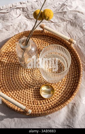 De ci-dessus des suppléments vitaminiques, y compris des capsules d'huile de poisson Oméga 3 et de la vitamine D3, soigneusement placés sur un plateau en rotin à côté d'un verre d'eau, sous doux Banque D'Images