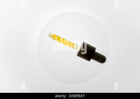 Gros plan d'une pipette en verre remplie d'huile jaune sur fond blanc. L'image met en évidence la transparence et la texture du liquide dans le cosme Banque D'Images