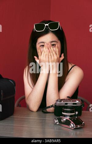 Une jeune femme asiatique est assise à une table avec les mains couvrant son visage et un appareil photo vintage à proximité. Elle porte des lunettes de soleil sur la tête et une tenue noire Banque D'Images