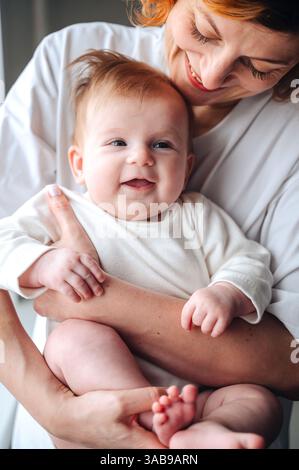 Mère aimante berce bébé nouveau-né souriant à la maison, les deux portant des vêtements blancs. L'expression joyeuse du bébé améliore la chaleur de leur partage Banque D'Images