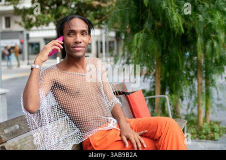 Un homme élégant est assis sur un banc, conversant sur un smartphone rose. Sa chemise en mesh avant-gardiste et son pantalon orange éclatant expriment confiance et fierté, Banque D'Images