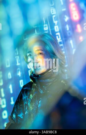 Une femme asiatique âgée expérimente l'intersection de la réalité et des mondes virtuels dans la métaverse. Entourée d'éléments numériques, elle explore de nouveaux t Banque D'Images