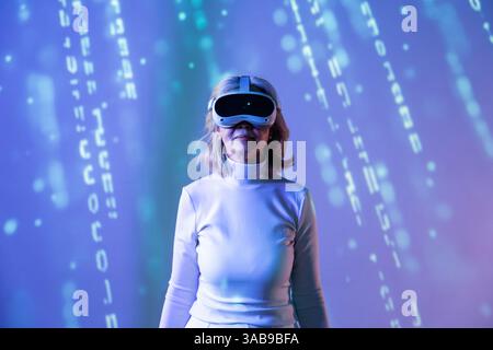 Une femme asiatique âgée, portant un casque VR, explore la métaverse, entourée de graphismes numériques. Elle expérimente la réalité virtuelle dans un style futuriste Banque D'Images
