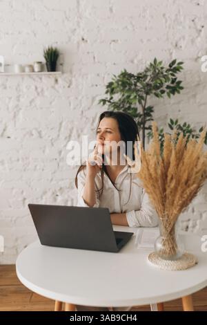 Un nutritionniste s'assoit soigneusement à un bureau blanc avec un ordinateur portable et de l'herbe de pampas dans un vase. L'espace lumineux est complété par un mur de briques et vert Banque D'Images