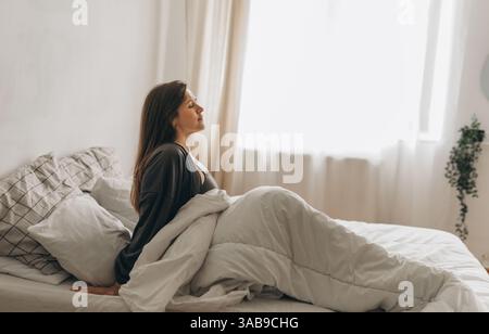 Une jeune femme est assise au lit les yeux fermés, enveloppée de couvertures blanches douces. Elle semble calme et détendue, profitant de la chaleur de la lumière du matin Banque D'Images