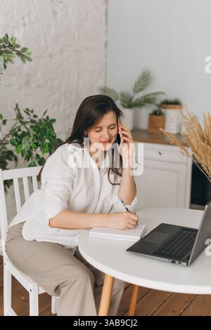 Un nutritionniste dans un bureau à domicile consulte un client par téléphone. Elle prend des notes sur un bloc-notes à côté d'un ordinateur portable, entourée d'un plan intérieur Banque D'Images
