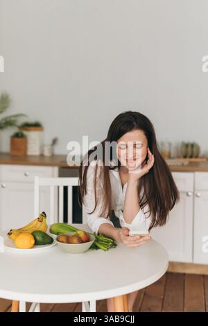 Un nutritionniste est dans une cuisine, souriant et utilisant un smartphone. Fruits et légumes frais sont sur la table. La scène transmet une alimentation saine et WEL Banque D'Images