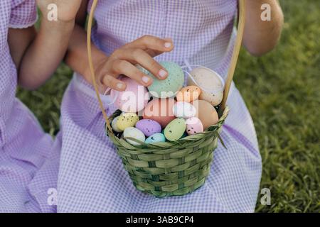 Un enfant tient un panier tissé vert rempli d'œufs de Pâques mouchetés et éclatants. La scène capture la joie et les couleurs des célébrations de Pâques sur une herbe Banque D'Images