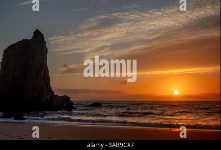 Vue panoramique sur une imposante formation rocheuse au coucher du soleil, avec le soleil projetant une lueur chaude sur les vagues de l'océan et la plage de sable, créant une image tranquille Banque D'Images