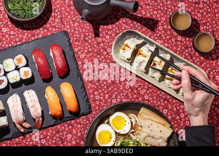 Composition artistique d'un festin japonais traditionnel, y compris des sushis, boulettes et ramen, avec une main visible à l'aide de baguettes, créant un naturel Banque D'Images