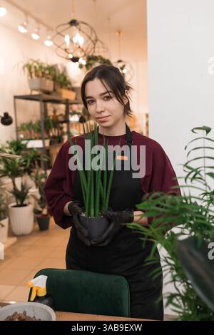 Une jeune femme dans un tablier tient une plante verte en pot avec des mains gantées dans un magasin de plantes intérieur. L'arrière-plan met en évidence des étagères de plantes luxuriantes et h. Banque D'Images