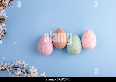Quatre œufs de Pâques mouchetés pastel sur fond bleu avec des branches de fleurs de cerisier et des pétales éparpillés. Concept de Pâques. Vue de dessus, pose à plat. Banque D'Images