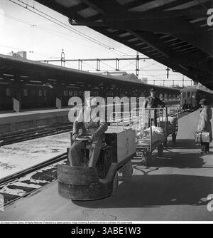 Une scène de gare des années 1950 Sur le quai, un homme conduit un petit véhicule électrique utilisé pour transporter des lettres et des colis par la poste. Attaché au véhicule est un chariot avec un autre homme se tient au-dessus du chariot, apparemment superviser le transport. À droite, une personne marche le long de la plate-forme, portant une mallette, éventuellement en chemin pour attraper un train. En arrière-plan, des wagons de train sont visibles à côté de fils électriques aériens, ce qui indique que la gare accueille des trains électriques. La plate-forme est abritée par une grande structure de toit, fournissant une couverture pour les passagers an Banque D'Images