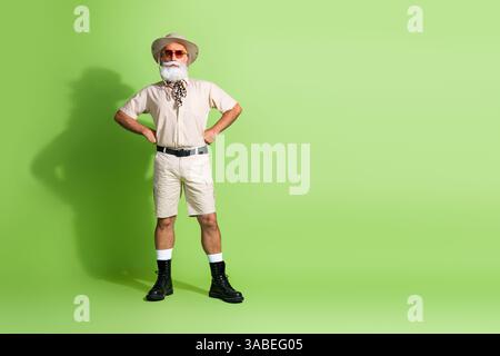 Explorateur âgé élégant dans une tenue de safari se tient en toute confiance sur un fond vert vibrant, embrassant l'aventure estivale et l'errance. Banque D'Images