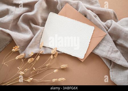 Vue aérienne rapprochée d'une carte blanche vierge et enveloppe sur une table avec une plante séchée décorative et un tissu de lin sur une table Banque D'Images
