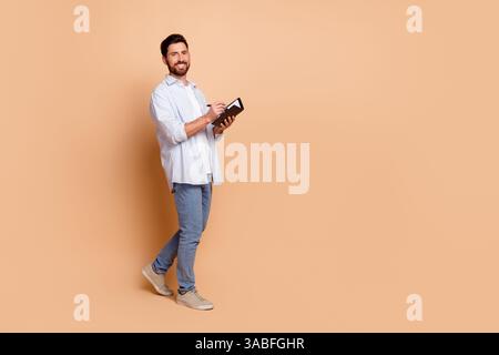 Un jeune homme décontracté tient une tablette tout en posant avec confiance sur un fond beige mettant en valeur le style et la personnalité Banque D'Images