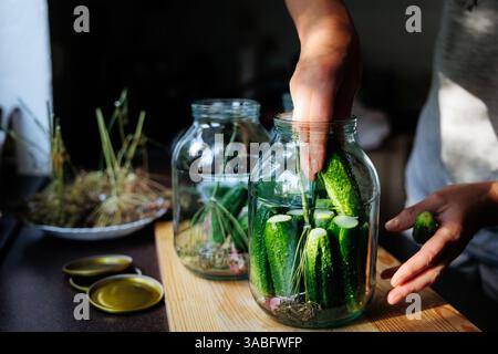 Femme mettant le concombre dans un pot de cornichon à table dans la cuisine Banque D'Images