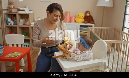 Mère et fille dans la chambre avec jouets et berceau, mère vérifiant le téléphone pendant que le tout-petit joue dans la chaise haute, intérieur rempli d'objets colorés et Banque D'Images