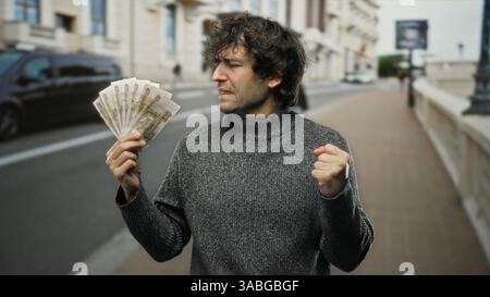Homme hispanique tenant des roubles russes dans la rue de la ville, mettant en valeur l'excitation et le succès dans un cadre urbain public. Banque D'Images
