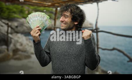 Homme hispanique excité tenant des roubles russes à la plage de bord de mer, célébrant en plein air avec un sourire joyeux au milieu de la nature et de l'océan en toile de fond, se sentant tr Banque D'Images
