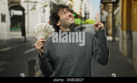Homme hispanique à l'extérieur dans la ville tenant des roubles russes tout en souriant et pointant vers le haut, suggérant le succès financier ou la richesse. Banque D'Images