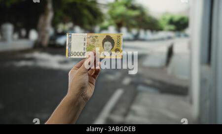 Homme tenant un billet de banque sud-coréen sur une rue de la ville à l'extérieur par une journée ensoleillée, mettant en valeur la monnaie dans un cadre urbain moderne avec un fond flou. Banque D'Images