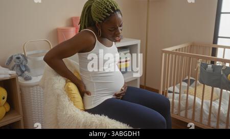 Femme avec des tresses éprouvant des maux de dos est assise sur un lit dans une chambre confortable près d'un berceau de bébé, dépeignant une atmosphère accueillante et nourrissante. Banque D'Images