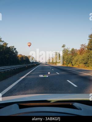 Point de vue : conduite avec régulateur de vitesse sur une autoroute vide alors que l'aérostat de ballon planait au-dessus. Arbres verts et arbustes sur les côtés de la route. Banque D'Images
