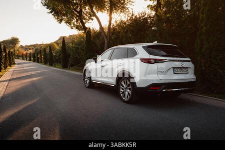 Mazda CX-9 blanc sur une route pavée de campagne bordée de cyprès. Vue de trois quarts arrière grand angle du VUS familial au coucher du soleil. Banque D'Images
