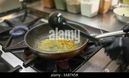 Chef professionnel portant des gants noirs en utilisant une louche pour mélanger les épices et le beurre dans une poêle sur une cuisinière à gaz professionnelle préparant un plat indien dans un comm Banque D'Images