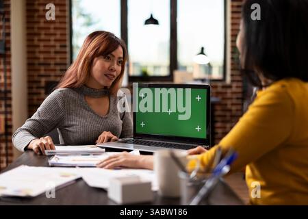 Ordinateur portable affichant l'écran vert comme femme d'affaires asiatique parle à son collègue au bureau, discutant des stratégies de marketing. Les collègues féminins partagent des plans de projet, en utilisant un appareil numérique avec modèle de maquette. Banque D'Images
