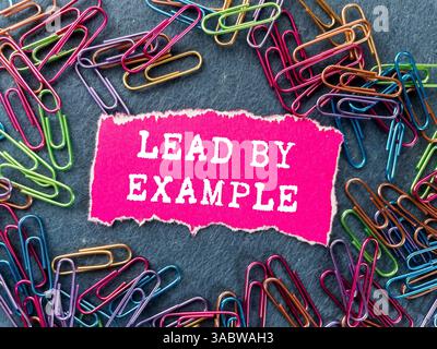 Dirigez par l'exemple un message écrit sur un morceau de papier rose déchiré sur fond gris. Symbole de la direction conceptuelle par exemple. Copier l'espace. Banque D'Images