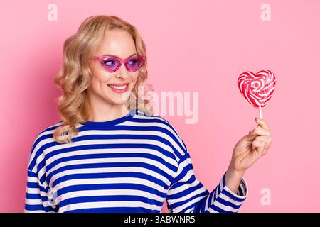 Charmante femme blonde dans un pull rayé élégant tenant un bonbon en forme de coeur, souriant joyeusement sur un fond rose joyeux Banque D'Images