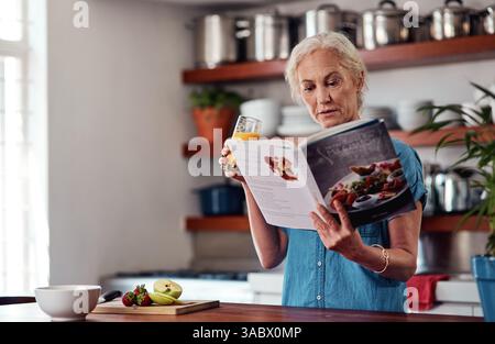 Femme mature, boisson et livre de recettes pour le régime, les ingrédients et lire pour les instructions dans la cuisine. Vegan, orange et cuisine pour la nourriture à la maison, femelle Banque D'Images