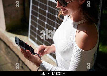 Femme connectant un smartphone à un panneau solaire photovoltaïque. Vue rognée. Intégration de l'énergie renouvelable durable dans la vie quotidienne, démonstration de l'utilisation pratique de l'énergie solaire pour les appareils de recharge. Banque D'Images