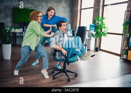Groupe de collègues s'amusant au bureau roulant sur une chaise, mettant en valeur le travail d'équipe et la camaraderie dans un environnement de travail moderne. Banque D'Images