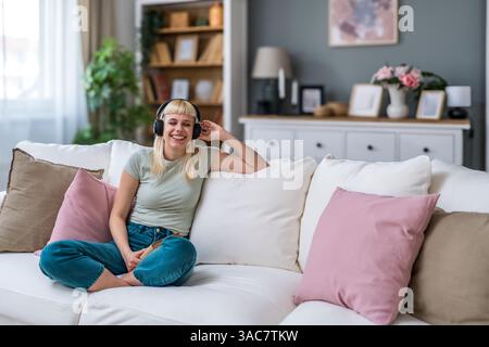 Jeune femme dans des salons de vêtements décontractés sur le canapé écoutant de la musique avec des écouteurs sans fil, lisant un livre. Il est temps de lire, amoureux du livre femme profitant w Banque D'Images