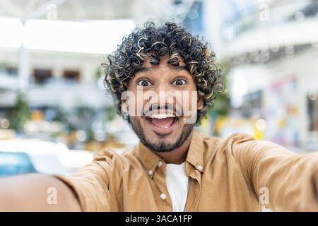 Un jeune homme aux cheveux bouclés prend un selfie, son visage éclairé de surprise et d'excitation. Sa bouche ouverte révèle des dents blanches, soulignant son bonheur. Banque D'Images