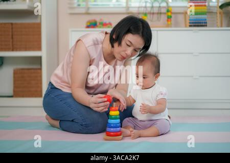 Une mère guidant son bébé pour empiler des anneaux en bois colorés sur un jouet de cheville montre le développement précoce des compétences. Banque D'Images
