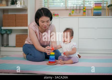 Une mère guidant son bébé pour empiler des anneaux en bois colorés sur un jouet de cheville montre le développement précoce des compétences. Banque D'Images