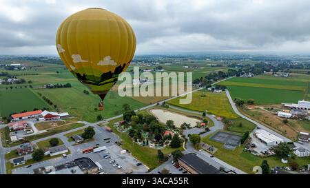 Une montgolfière dynamique flotte gracieusement au-dessus d'un paysage rural pittoresque rempli de champs verts et de bâtiments dispersés. Le ciel nuageux ajoute une atmosphère sereine à la scène. Banque D'Images