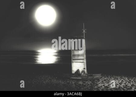 Une photographie sereine en noir et blanc représentant une tour de sauveteurs et un bateau de sauvetage au premier plan, avec la plage et la mer reflétant la pleine lune. Banque D'Images
