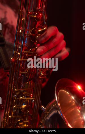 Gros plan vertical d'un saxophoniste jouant de la musique jazz enchanteresse en appuyant sur des touches d'un instrument brillant avec des doigts agiles, tout en jouant sur scène sous un éclairage rouge Banque D'Images