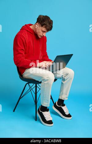 Jeune homme concentré avec des écouteurs travaillant sur un ordinateur portable tout en étant assis sur une chaise sur fond bleu de studio. Concept de télétravail, ventes. Banque D'Images