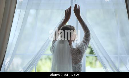 Vue arrière d'une mariée ouvrant gracieusement les rideaux tout en portant une robe de mariée élégante et voile sur son jour de mariage spécial Banque D'Images