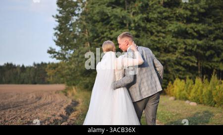 Couple de jeunes mariés se promenant ensemble, se tenant la main dans une prairie ensoleillée, partageant le moment intime du jour du mariage Banque D'Images