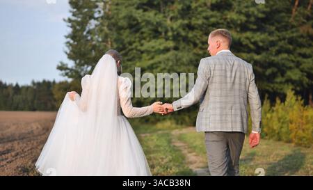 Couple de jeunes mariés marchant main dans la main le long du chemin extérieur, partageant un moment intime après la cérémonie de mariage Banque D'Images