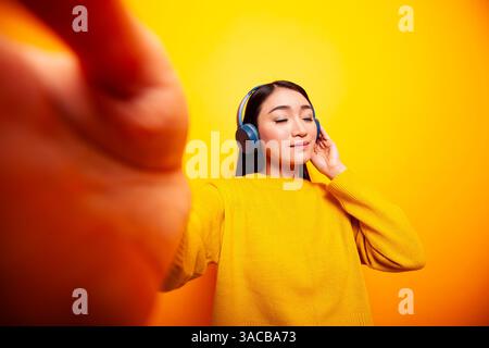 Vlogger asiatique debout sur fond isolé et écoutant de la musique. Jeune femme avec des écouteurs, enregistrant un vlog quotidien et tenant une caméra, avec une expression détendue devant un fond jaune. Banque D'Images